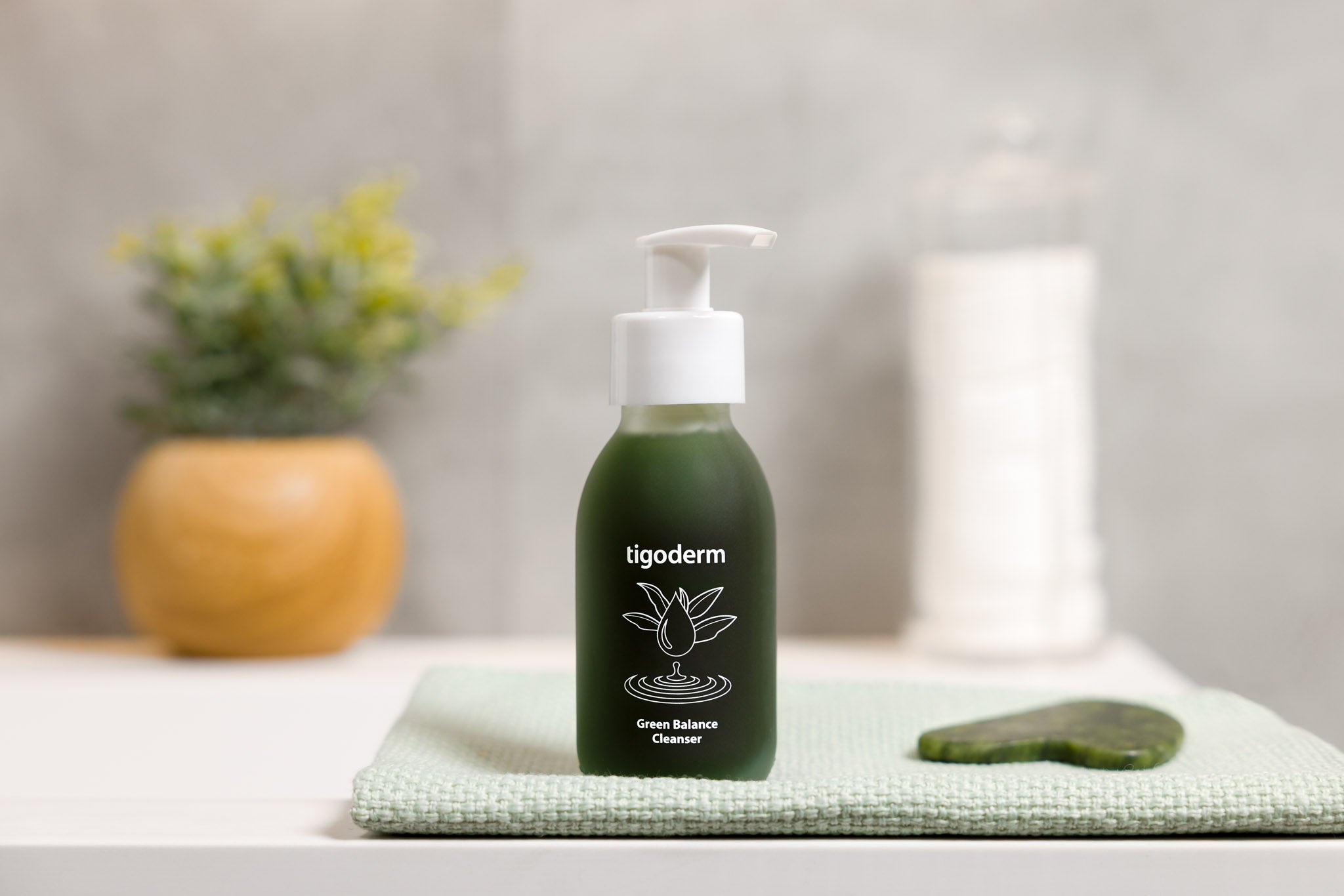 tigoderm Green Balance Waschgel im Glasflakon mit Pumpkopf auf einem Handtuch neben einem Gua Sha Stein.