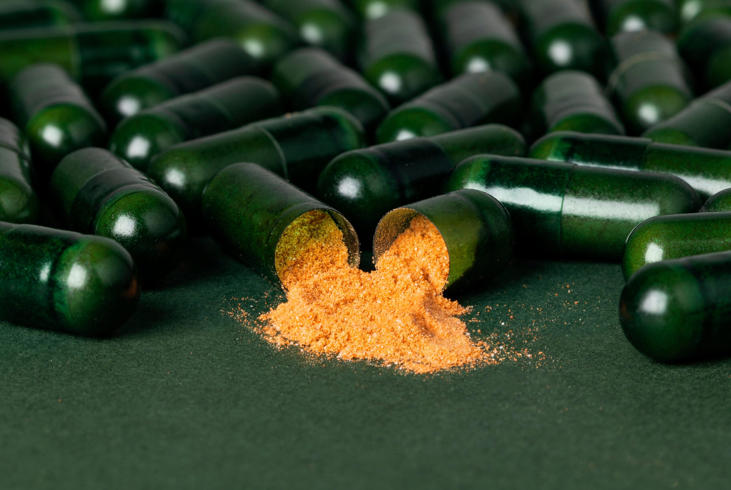 Geöffnete tigovit Grüntee-Extrakt Kapsel mit sichtbarem EGCG-Pulver vor einem Hintergrund aus dunkelgrünen Kapseln.