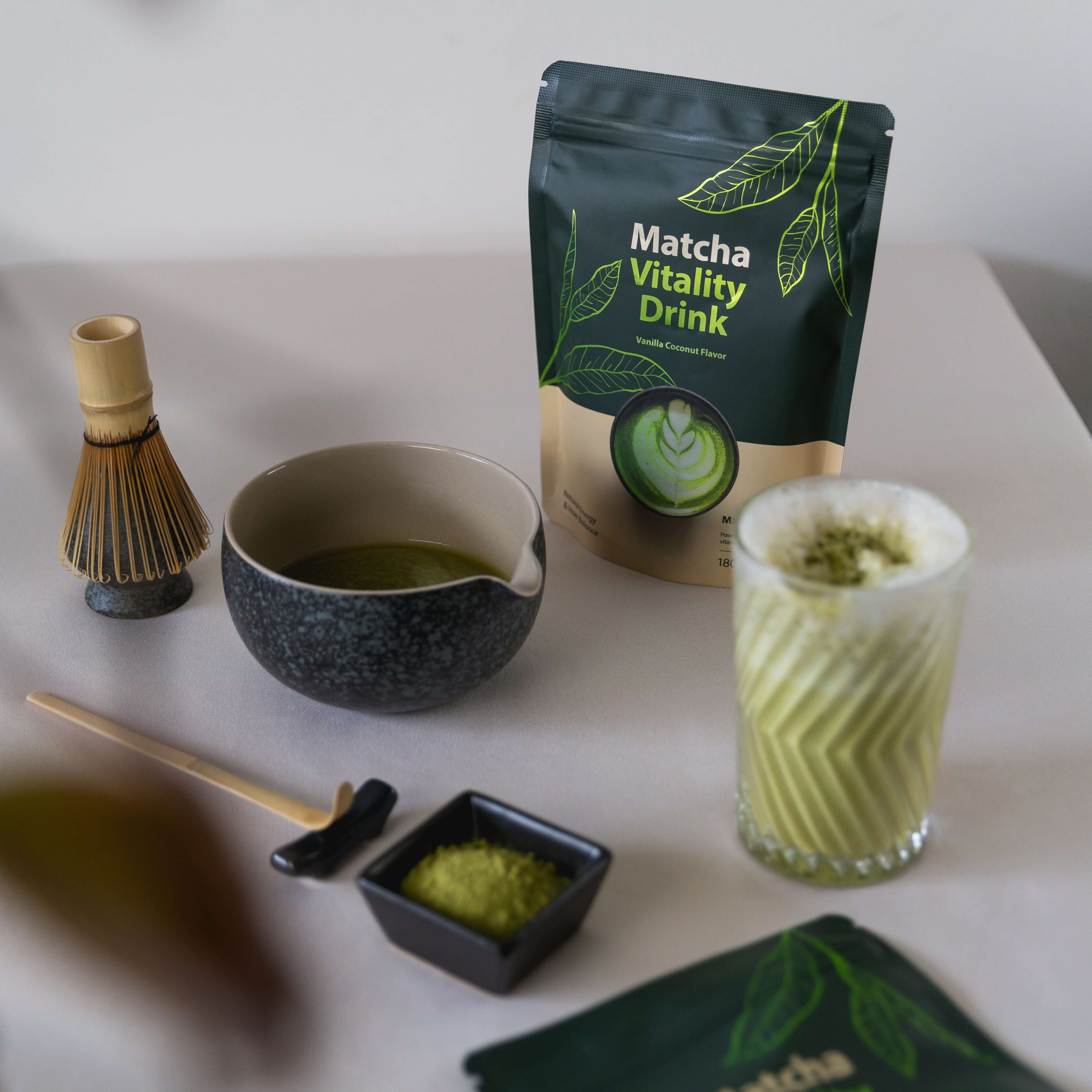 Matcha Vitality Drink mit Kokos-Vanille-Geschmack neben Bambusbesen, Schale und zubereitetem Matcha Latte.